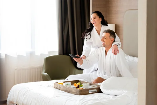 Novia sonriente en albornoz abrazando novio guapo con controlador remoto en el hotel - foto de stock