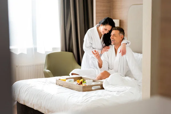 Smiling girlfriend in bathrobe hugging handsome boyfriend in hotel — Stock Photo