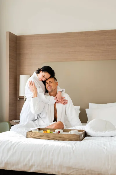 Novia sonriente en albornoz abrazando novio y mirando bandeja con comida en el hotel - foto de stock