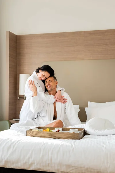 Petite amie souriante en peignoir étreignant petit ami et regardant plateau avec de la nourriture à l'hôtel — Photo de stock