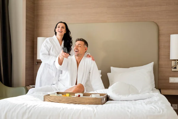Novia sonriente en albornoz abrazando novio guapo con controlador remoto en el hotel - foto de stock