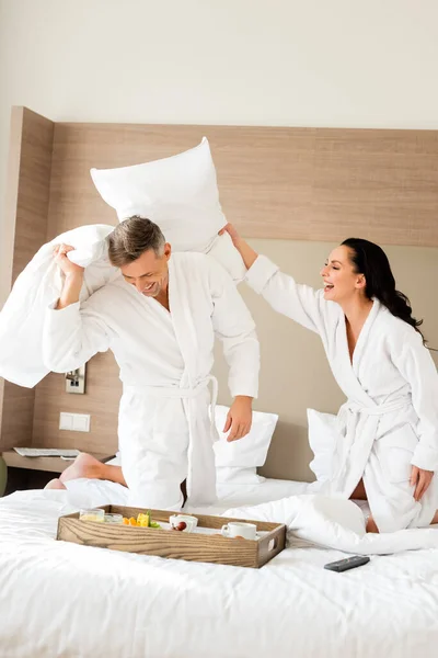 Sorrindo namorado e namorada em roupões de banho lutando com travesseiros no hotel — Fotografia de Stock