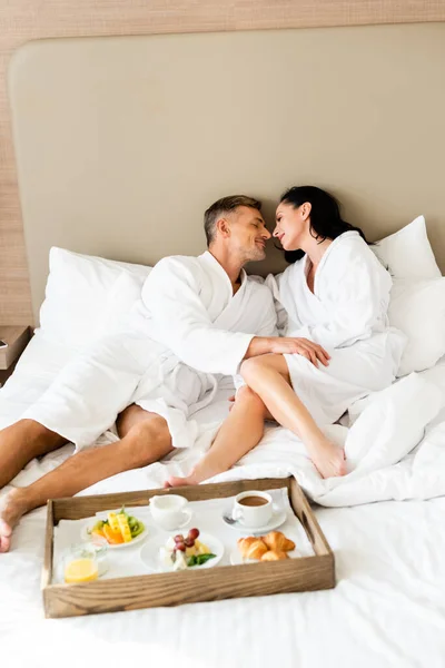 Novio y novia en albornoces besándose cerca de bandeja con comida en el hotel - foto de stock