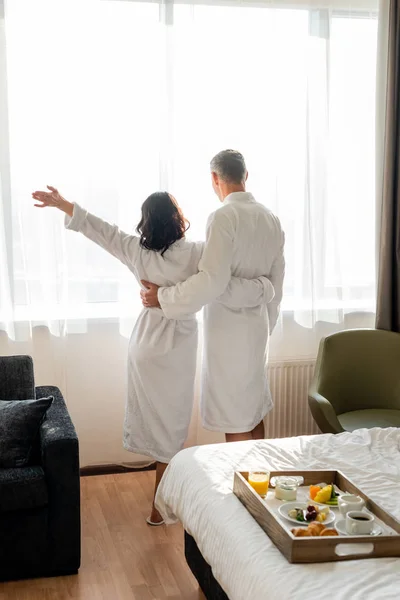 Back view of boyfriend and girlfriend in bathrobes hugging and looking trough window in hotel — Stock Photo
