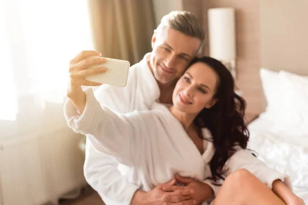 Selective focus of boyfriend hugging smiling girlfriend in bathrobe and she taking selfie in hotel — Stock Photo