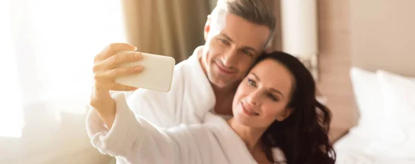 Plano panorámico de novio abrazando novia sonriente en albornoz y ella tomando selfie en el hotel - foto de stock