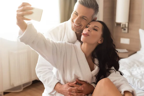 Novio abrazando sonriente novia en albornoz y ella tomando selfie y sobresaliendo lengua en el hotel - foto de stock