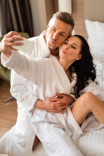 Boyfriend hugging girlfriend in bathrobe and she taking selfie and showing kiss in hotel — Stock Photo