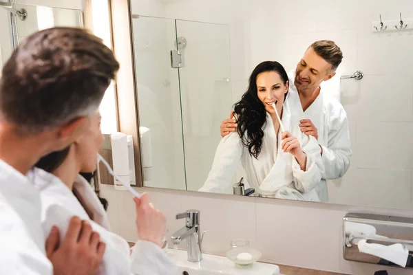 Selective focus of boyfriend hugging smiling girlfriend and she brushing teeth in hotel — Stock Photo