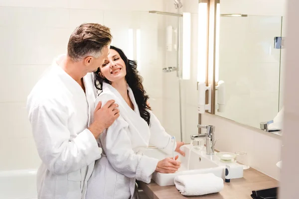 Freund umarmt lächelnde Freundin im Bademantel mit Zahnbürste im Hotel — Stockfoto
