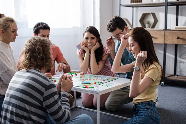 KYIV, UCRANIA - 27 de enero de 2020: chica emocionada mostrando el gesto ganador mientras juega al juego del monopolio con amigos sonrientes - foto de stock