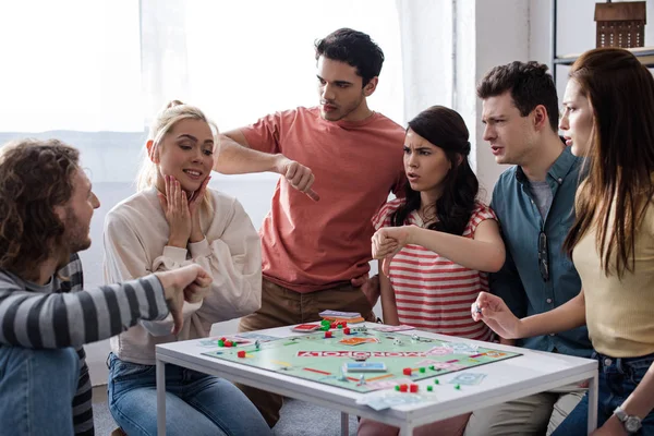 KYIV, UKRAINE - JANUARY 27, 2020: happy girl sitting near displeased friends showing thumbs down while playing monopoly game — Stock Photo