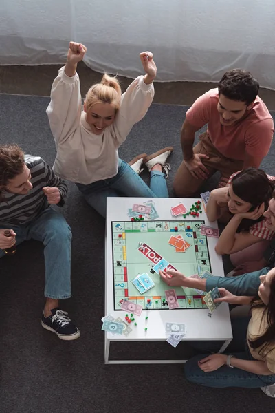 KYIV, UCRANIA - 27 de enero de 2020: vista aérea de la chica emocionada mostrando el gesto ganador mientras juega al juego del monopolio con amigos sonrientes - foto de stock