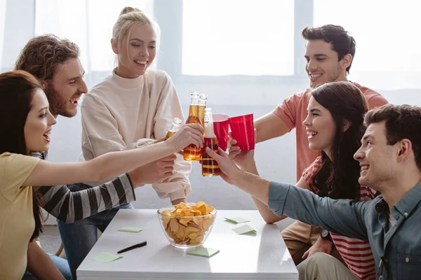 Amis joyeux cliquetis avec des bouteilles et des tasses en plastique tout en jouant jeu de nom à la maison — Photo de stock