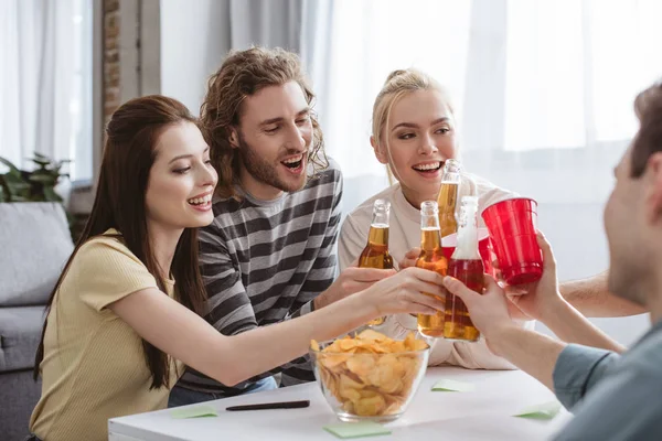 Amis heureux cliquetis avec des bouteilles et des tasses tout en jouant jeu de nom à la maison — Photo de stock