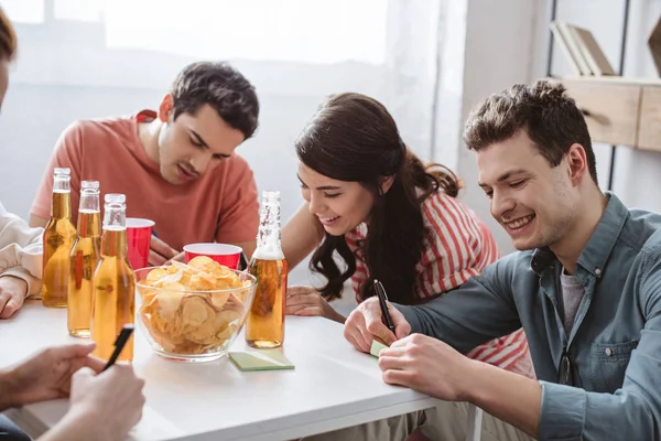 Amis joyeux écrivant sur des notes collantes tout en jouant jeu de nom à table avec des boissons et des jetons — Photo de stock