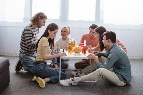 Cheerful friends sitting on floor at table with drinks and chips while playing name game — Stock Photo