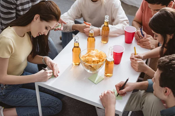 Vista de ángulo alto de amigos que escriben en notas adhesivas mientras juegan juego de nombre en la mesa con bebidas y patatas fritas - foto de stock