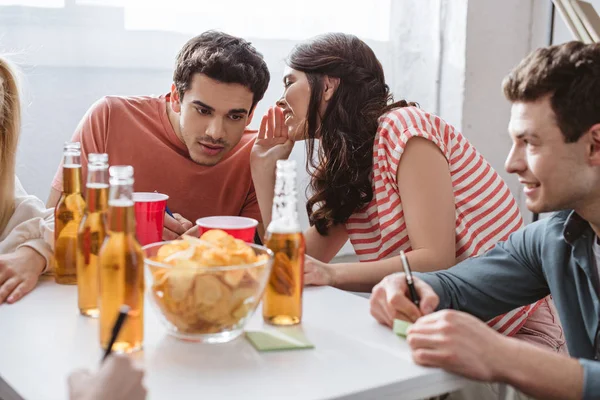 Girl whispering at attentive guy while playing name game with smiling friends — Stock Photo