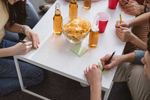 Обрезанный вид друзей, пишущих на липких нотах, играя в именную игру за столом с напитками и чипсами — стоковое фото