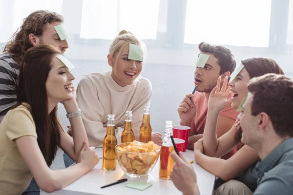 Amigos sorridentes com notas pegajosas na testa jogando nome jogo à mesa com bebidas e batatas fritas — Fotografia de Stock