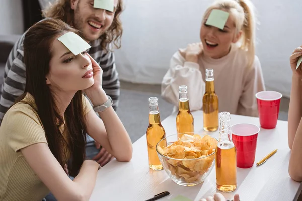Thoughtful girl playing name game with cheerful friends with sticky notes on foreheads — Stock Photo