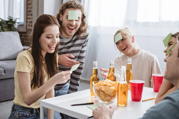 Cheerful girl looking at sticky note while playing name game with happy friends — Stock Photo