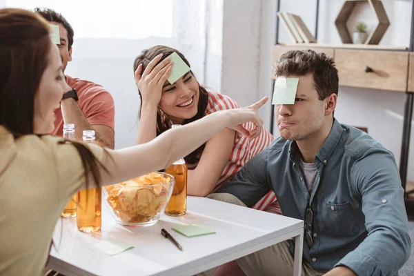 Selective focus of girl pointing with hand at man with sticky note on forehead while playing name game with friends — Stock Photo