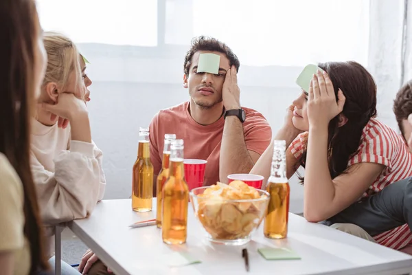 Amigos reflexivos con notas pegajosas en la frente jugando juego de nombre en la mesa con bebidas y patatas fritas - foto de stock