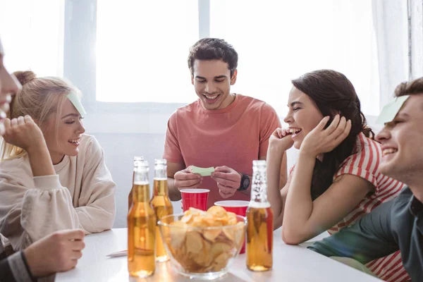 Hombre alegre mirando nota pegajosa mientras juega juego de nombre con amigos felices - foto de stock