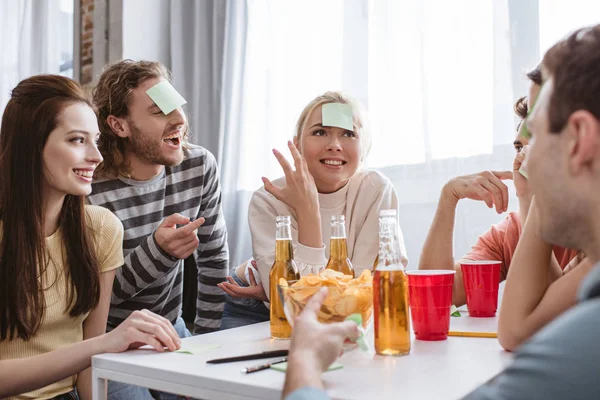 Cheerful friends with sticky notes on foreheads playing name game at table with beer and chips — Stock Photo