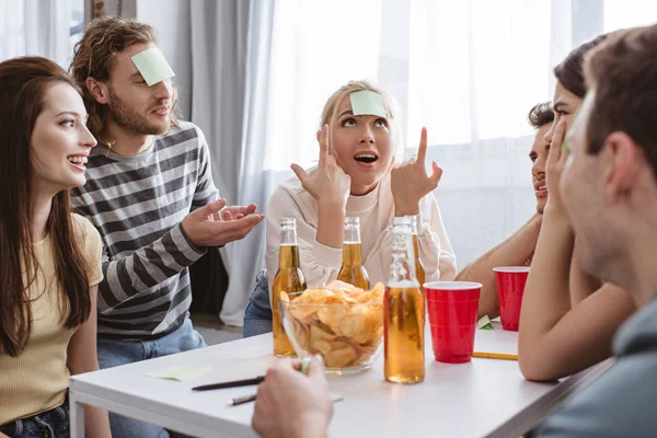 Alegre chica apuntando con el dedo a la nota pegajosa en su frente mientras juega juego nombre con amigos - foto de stock