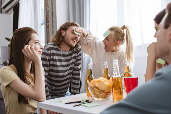 Chica feliz poner nota pegajosa en la frente del hombre sonriente mientras juega juego de nombre con amigos - foto de stock