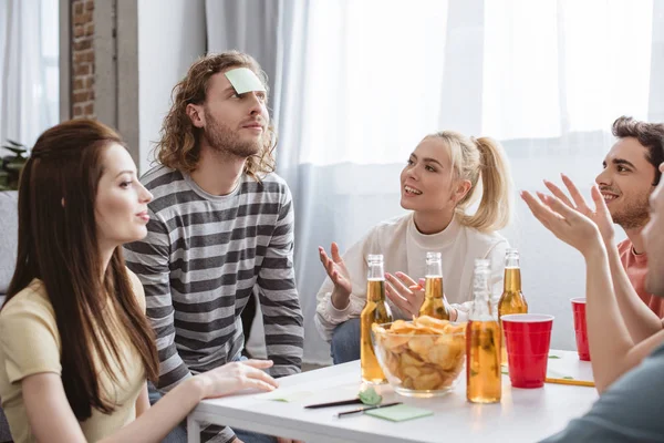 Amis souriants regardant l'homme avec une note collante sur le front tout en jouant jeu de nom — Photo de stock