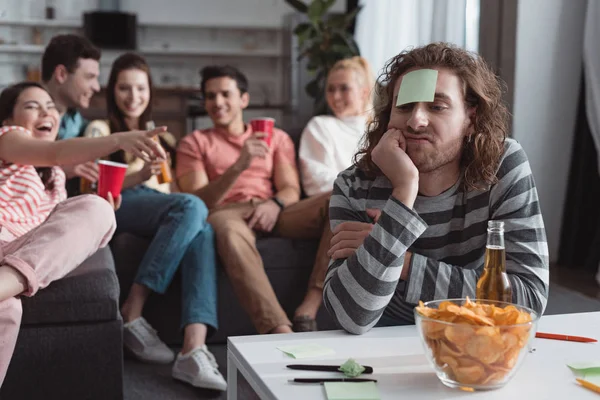 Joyeuse fille pointant avec la main à l'homme ennuyé avec une note collante sur le front tout en jouant jeu de nom avec des amis — Photo de stock