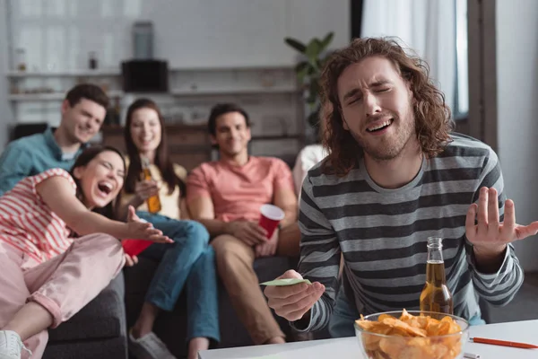 Selektiver Fokus des unzufriedenen Mannes, der eine klebrige Note hält, während er mit fröhlichen Freunden ein Namensspiel spielt — Stockfoto