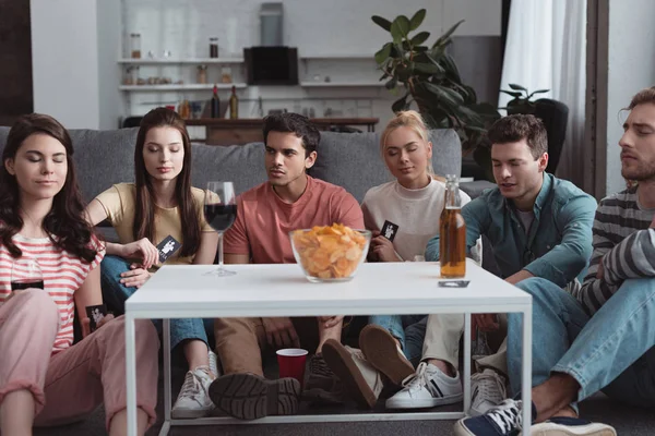 QUIIV, UCRÂNIA - JANEIRO 27, 2020: amigos atenciosos jogando jogo de máfia enquanto se sentam perto da mesa com batatas fritas e bebidas — Fotografia de Stock