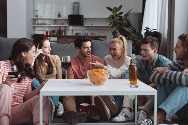 Excited girl pointing with hand while sitting at table with chips and drinks near cheerful friends — Stock Photo