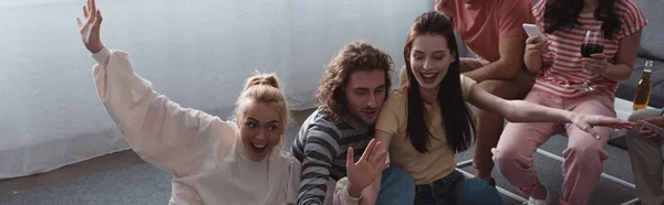 Panoramic shot of cheerful girl waving hand while sitting with friends at home — Stock Photo