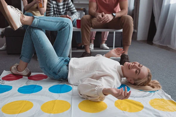 KYIV, UCRANIA - 27 de enero de 2020: niña alegre sonriendo mientras está acostada en la alfombra de juego de Twister cerca de amigos - foto de stock