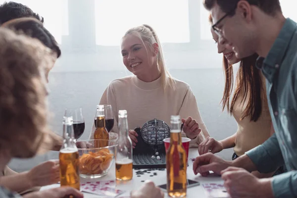 KYIV, UCRANIA - 27 de enero de 2020: amigos alegres jugando al juego de lotería mientras están sentados en la mesa con bebidas y papas fritas - foto de stock