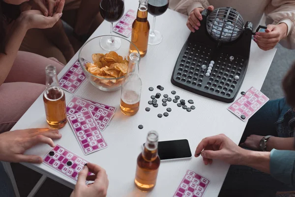 KYIV, UCRANIA - 27 de enero de 2020: vista recortada de amigos sentados en la mesa con dispensador de bolas de lotería, tarjetas, chips de cobertura y teléfono inteligente con pantalla en blanco - foto de stock