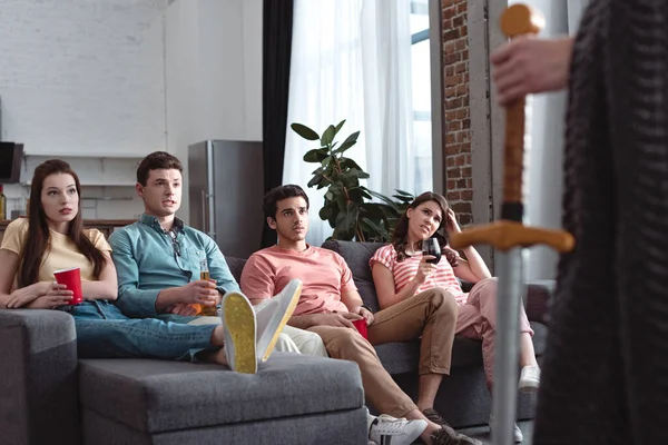 Partial view of man in fairy king costume standing near serious friends sitting on sofa — Stock Photo