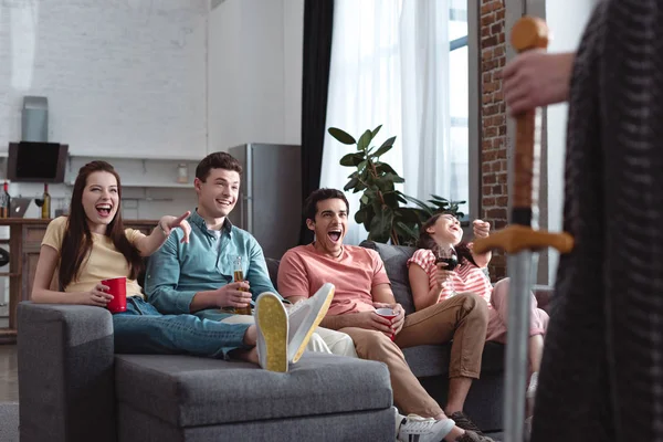 Частичный вид человека в сказочном костюме короля, стоящего рядом со смеющимися друзьями, сидящими на диване — стоковое фото