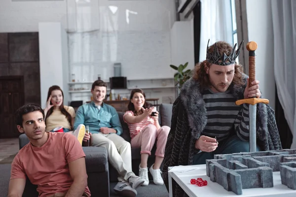 Kiew, Ukraine - 27. Januar 2020: Nachdenklicher Mann im Kostüm eines Königs beim Labyrinthspiel vor lächelnden Freunden — Stockfoto