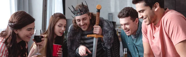 Panoramic shot of man in costume of king pointing with finger near cheerful friends — Stock Photo