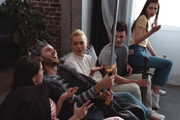 Cheerful man in costume of king holding toy sword while sitting on sofa near friends — Stock Photo