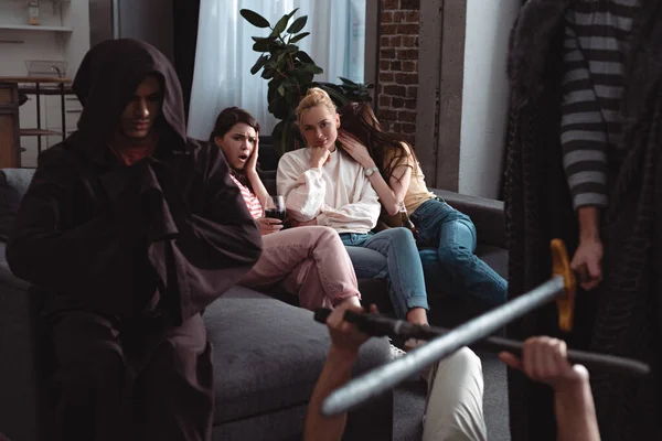 Cropped view of men fighting with toy swords near guy in costume of monk while worried girls sitting on sofa — Stock Photo