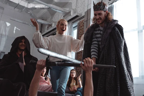 Men in costume of king fighting with knight with toy swords near friends — Stock Photo