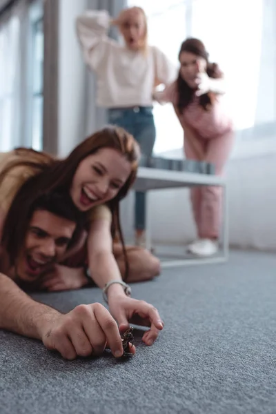 Selective focus of cheerful girl trying to take toy figurine from guy lying on floor and excited girls standing on background — Stock Photo